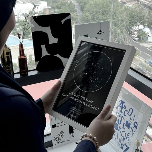 Person holding a birthday custom star map print with the quote "Look at the stars that witnessed your birth," framed and displayed by a window.