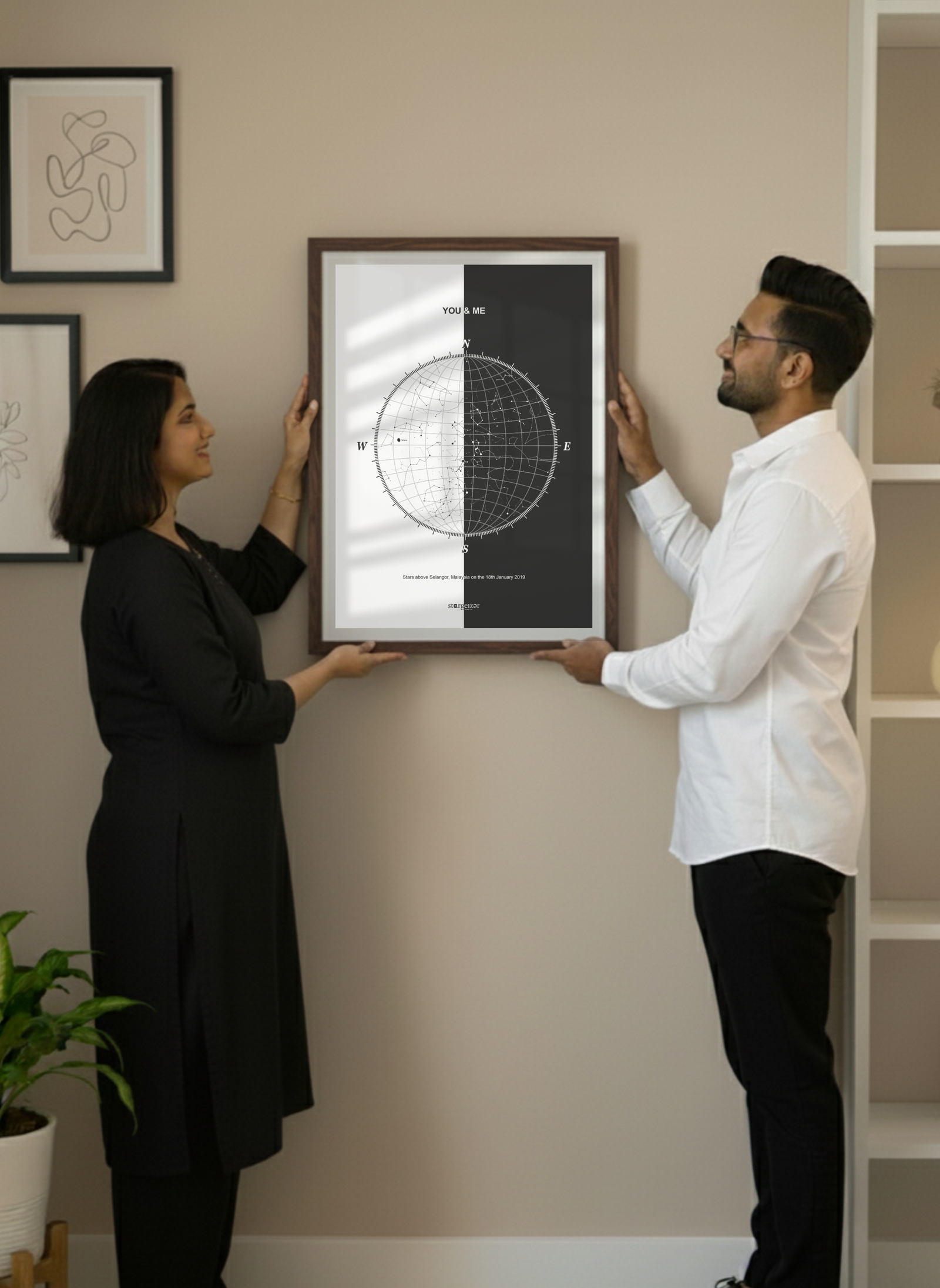 A couple in black and white outfits holding up a large framed custom star map artwork against a beige coded wall, with decorative frames and shelves in the background.