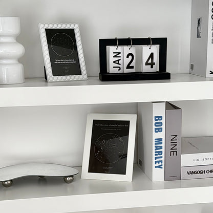White shelf with framed star map, decorative items including a vase and a calendar.