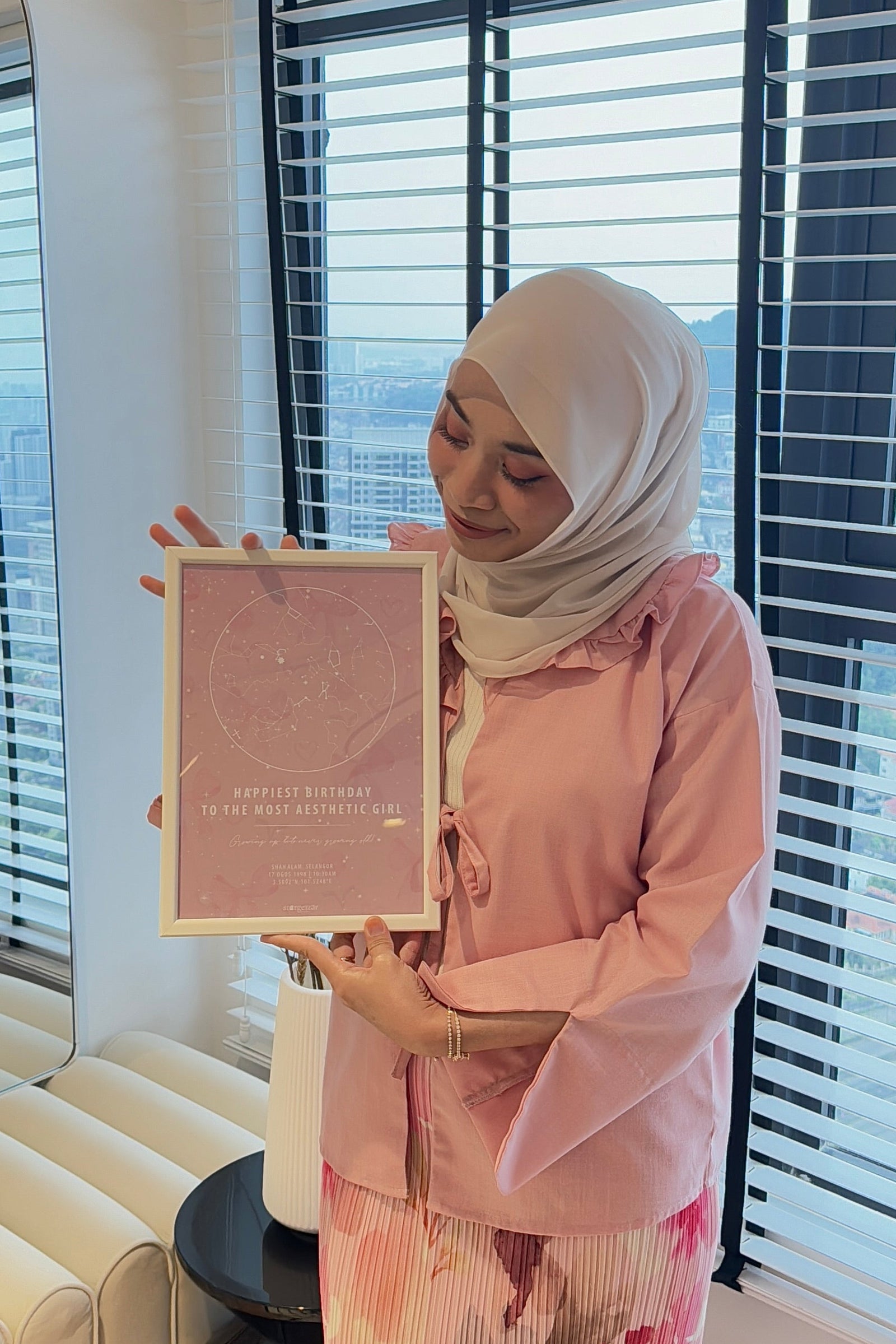 Woman in a light pink outfit holding a pastel pink star map by the window, natural daylight highlighting the personalized gift from Stargeizer.