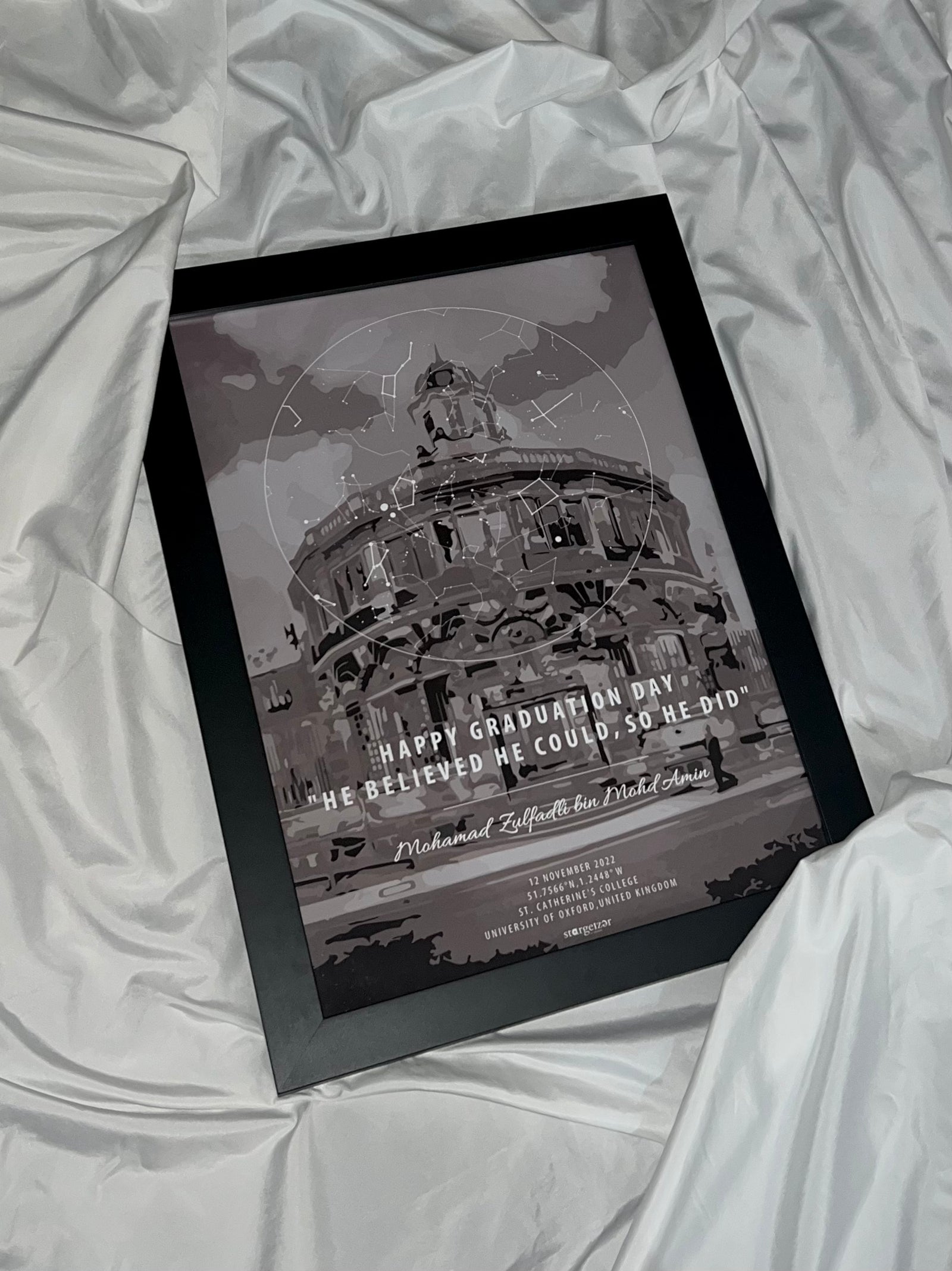 Framed custom star map with illustrated university building and graduation quote, displayed on silky white fabric.