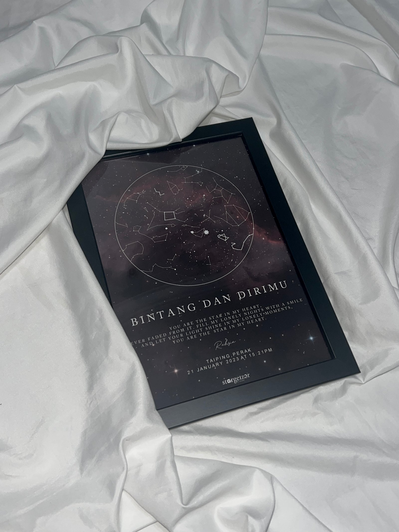 Framed custom star map with deep galaxy design and personalized Malay dedication, placed on white draped fabric.