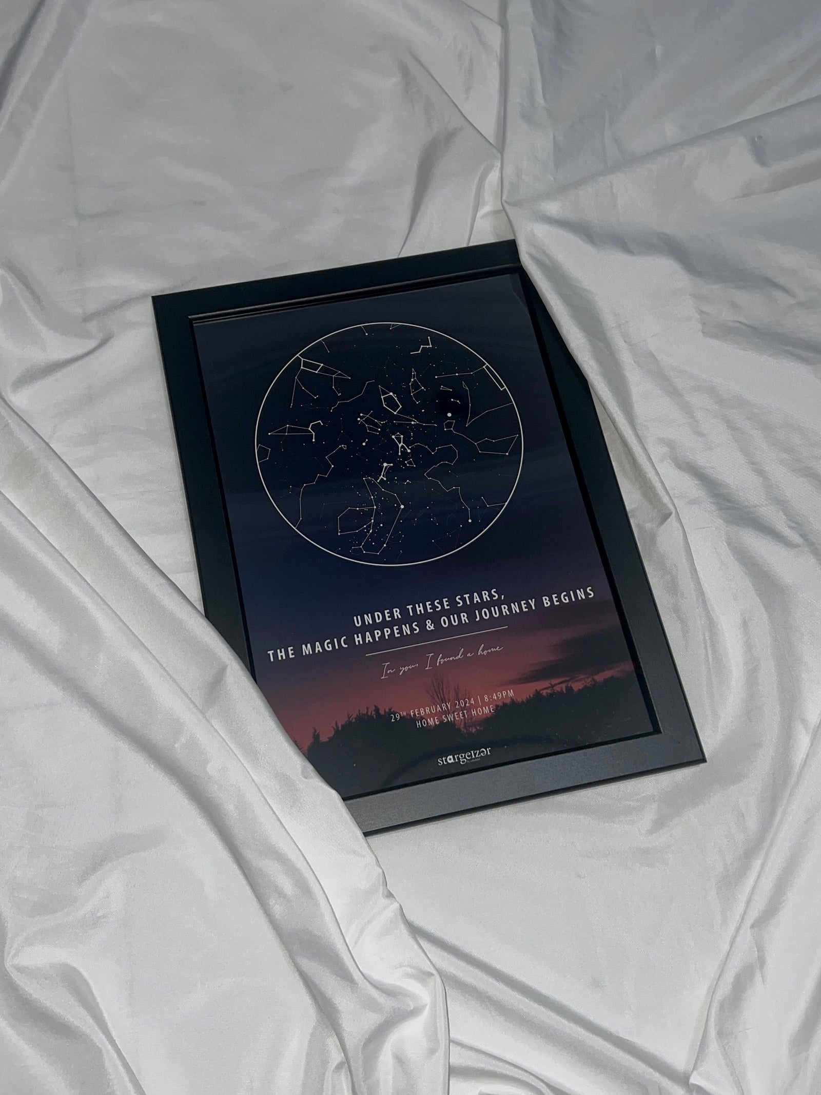 Framed star map with twilight sky background and personalized couple’s message, displayed on silky white sheet.