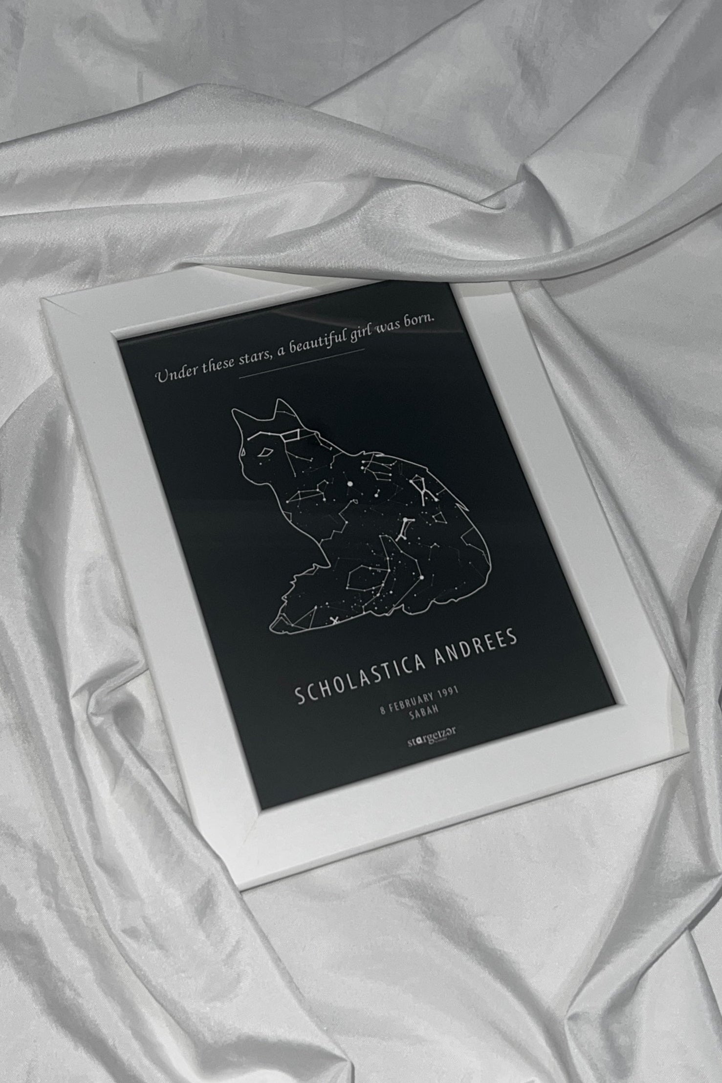 Framed star map in the shape of a cat with personalized name and date engraving on a silky white fabric background.
