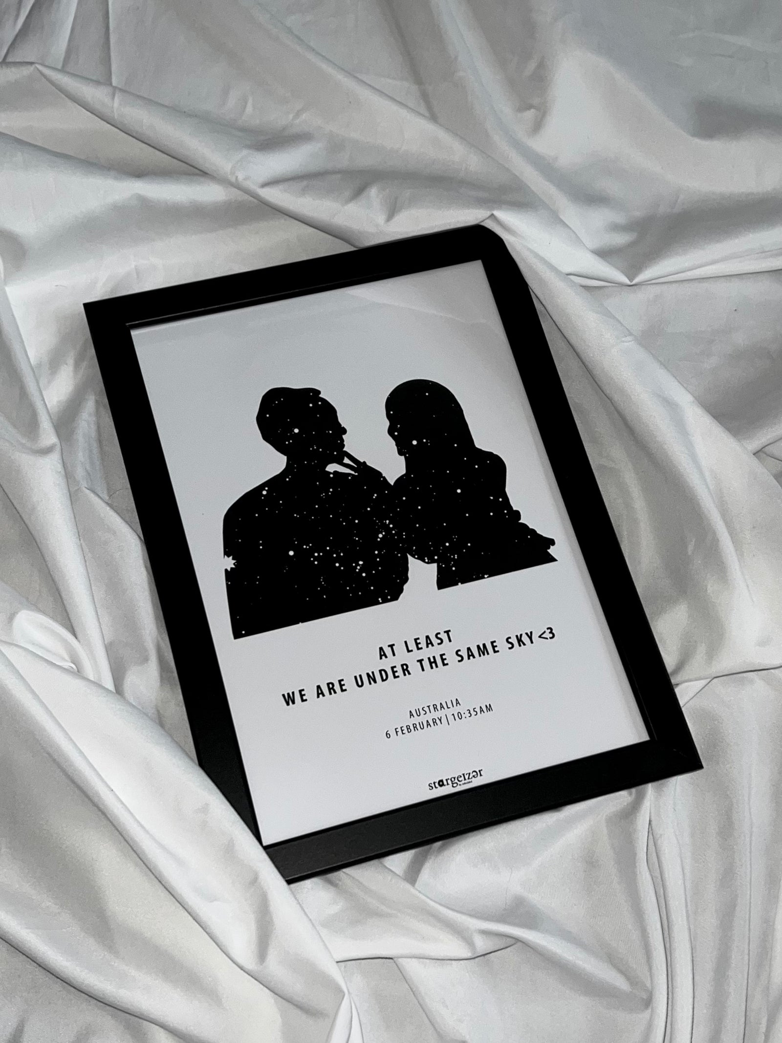 Black framed custom star map of a couple silhouette under the stars with message and date, displayed on silky white fabric.