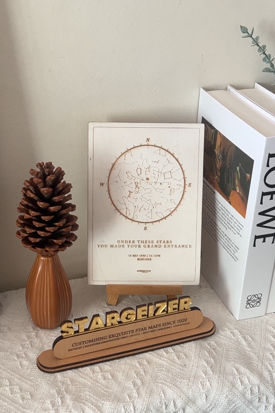 Minimalist beige wooden engrave  star map displayed on a wooden stand beside a pine cone and Loewe book, styled in a cozy neutral setting by Stargeizer.